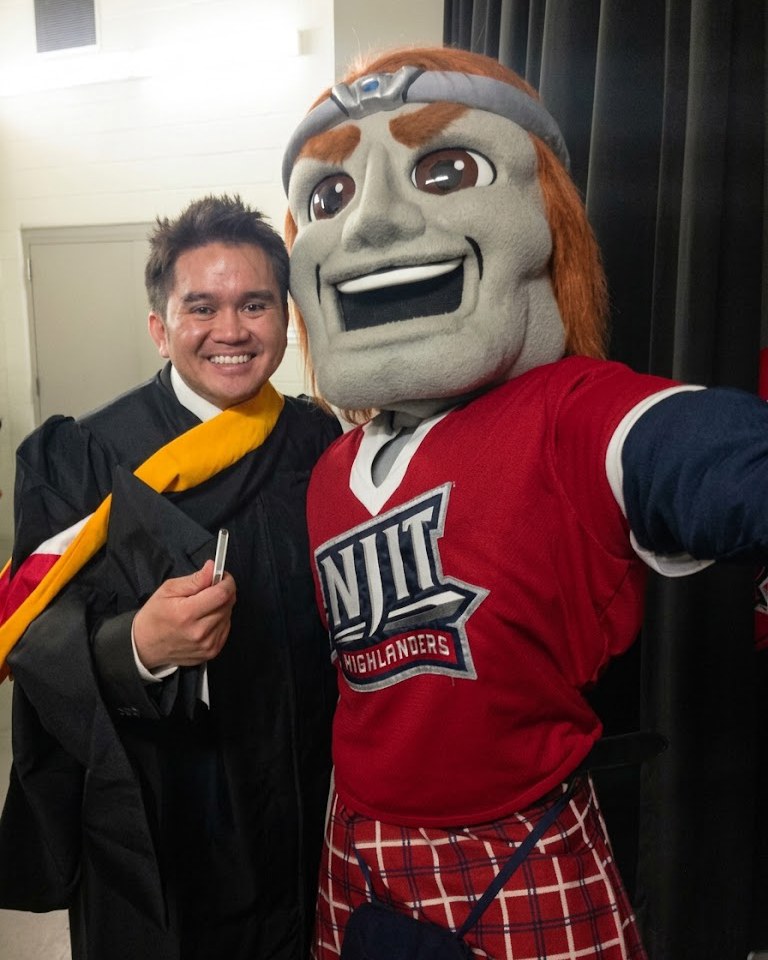 Ivan Balasi in graduation robes, smiling beside the NJIT Highlanders mascot