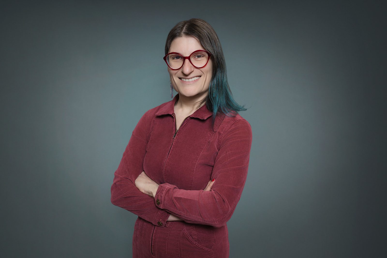 Studio headshot of a smiling woman in red glasses and a burgundy jacket, arms crossed